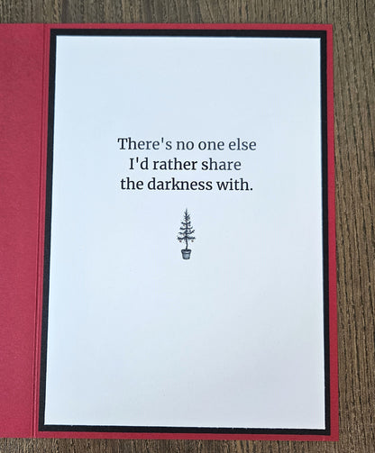 A handmade greeting card with a white center panel and red border, placed on a wood surface. The card reads, 'There's no one else I'd rather share the darkness with.