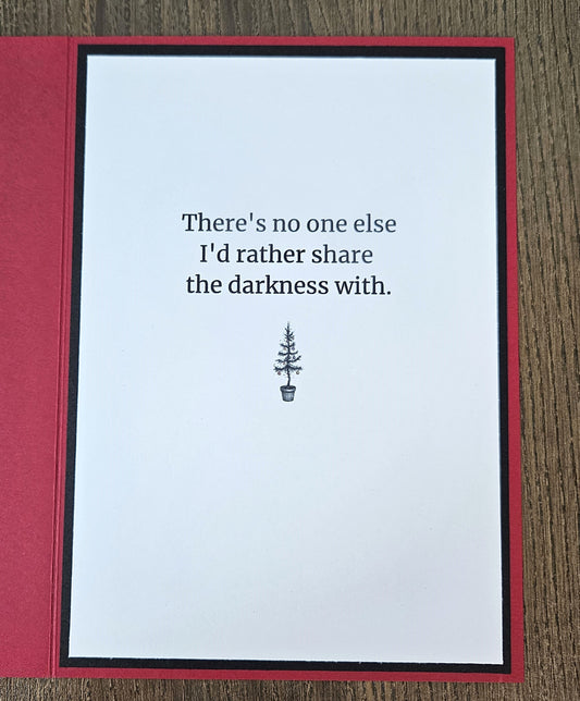 A handmade greeting card with a white center panel and red border, placed on a wood surface. The card reads, 'There's no one else I'd rather share the darkness with.
