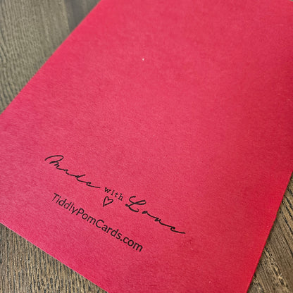 A close-up of a bright red textured card or envelope corner printed with the words 'Made with Love' and 'TidilyPomCards.com' in black script, placed on a wood surface.