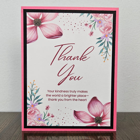 A handmade thank you card with a pink border, featuring watercolor pink flowers, green leaves, and the words 'Thank You' in cursive, standing on a dark wooden surface against a light gray wall.