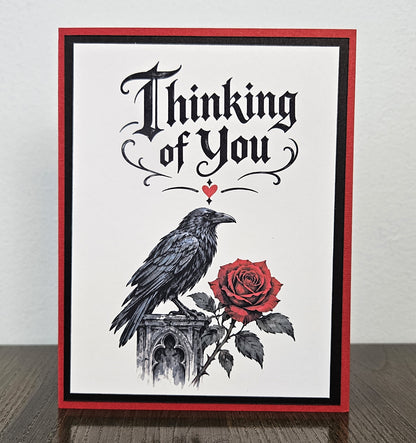 Raven and red rose Thinking of You card lying flat with a white envelope behind it.