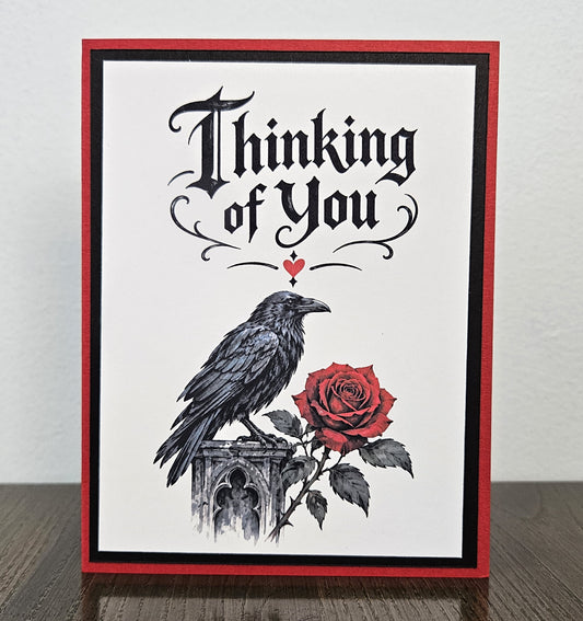 Raven and red rose Thinking of You card lying flat with a white envelope behind it.