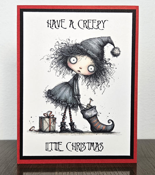 A handmade greeting card featuring a creepy, gothic-style illustration of a girl with wild hair wearing a Santa hat and holding a Christmas stocking, with the text 'HAVE A CREEPY LITTLE CHRISTMAS'. The card has a red border, a black inner border, and a white center, shown standing on a dark wooden surface against a light gray wall.