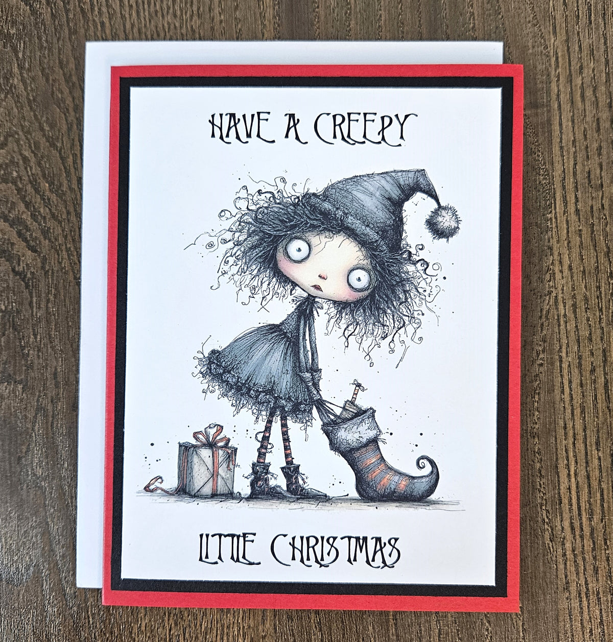 A greeting card featuring a gothic horror Christmas illustration, photographed on a wooden surface. The card has a white center with a red border, and the artwork shows a creepy doll-like figure with wild hair, a pointed hat, a stocking, and wrapped gifts.