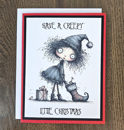 A greeting card featuring a gothic horror Christmas illustration, photographed on a wooden surface. The card has a white center with a red border, and the artwork shows a creepy doll-like figure with wild hair, a pointed hat, a stocking, and wrapped gifts.