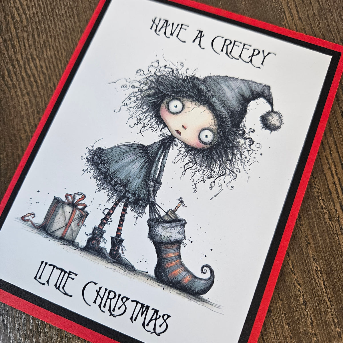 A greeting card featuring a gothic horror Christmas illustration, photographed on a wooden surface. The card has a white center with a red border, and the artwork shows a creepy doll-like figure with wild hair, a pointed hat, a stocking, and wrapped gifts.