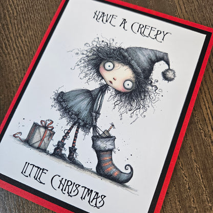 A greeting card featuring a gothic horror Christmas illustration, photographed on a wooden surface. The card has a white center with a red border, and the artwork shows a creepy doll-like figure with wild hair, a pointed hat, a stocking, and wrapped gifts.