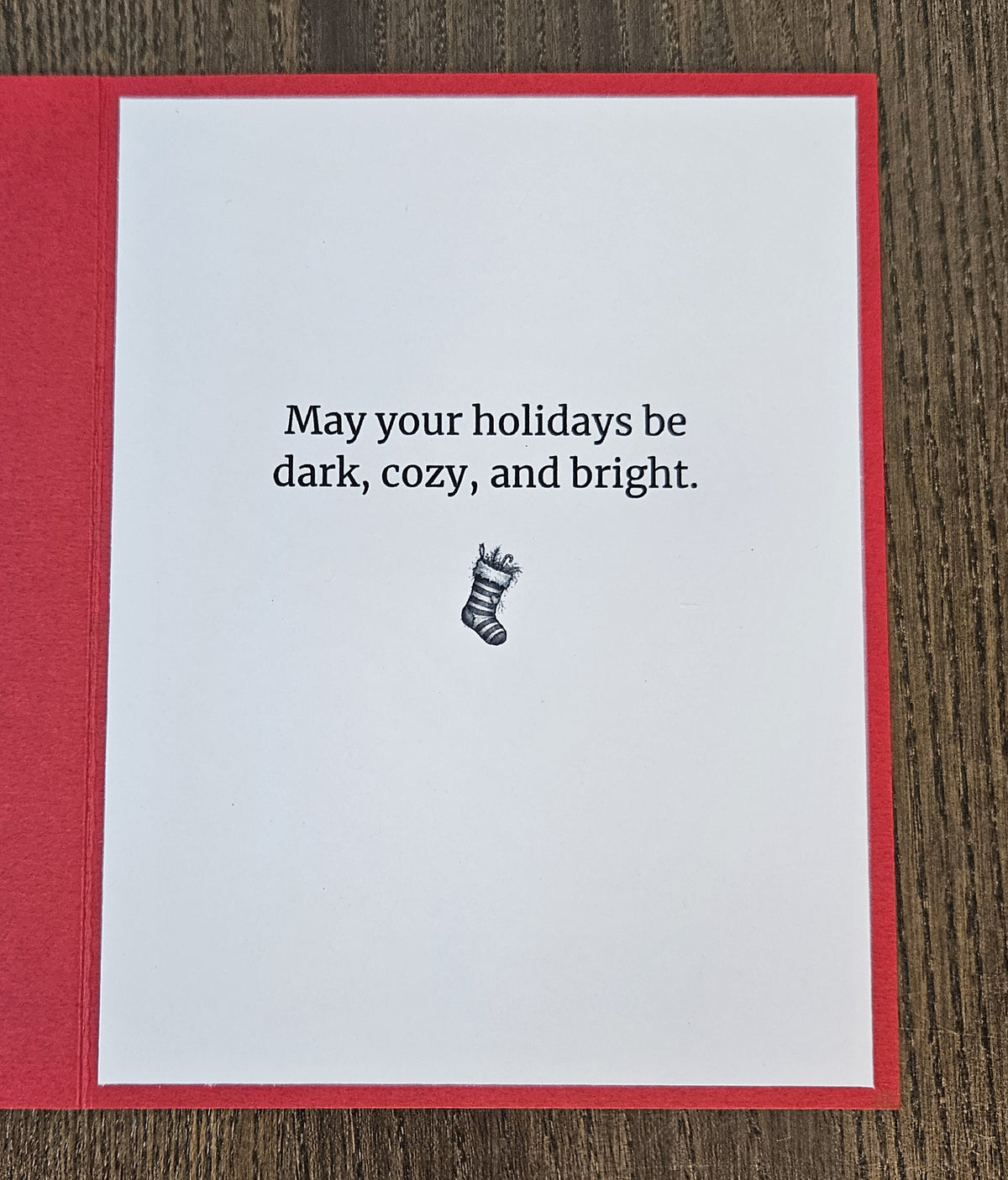 A greeting card with a red border and white center, placed on a dark wood surface. The card reads, 'May your holidays be dark, cozy, and bright.