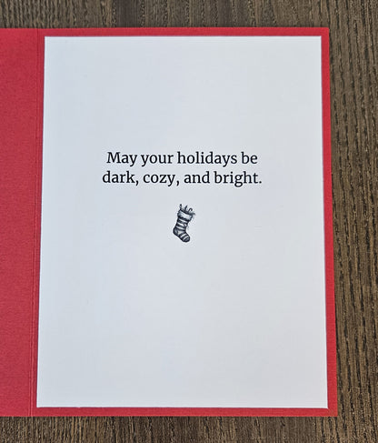 A greeting card with a red border and white center, placed on a dark wood surface. The card reads, 'May your holidays be dark, cozy, and bright.