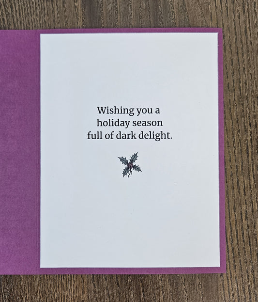 Handmade gothic Christmas card reading Deck the Halls with Dread and Holly, featuring a stitched-smile character with presents, framed in black and purple.