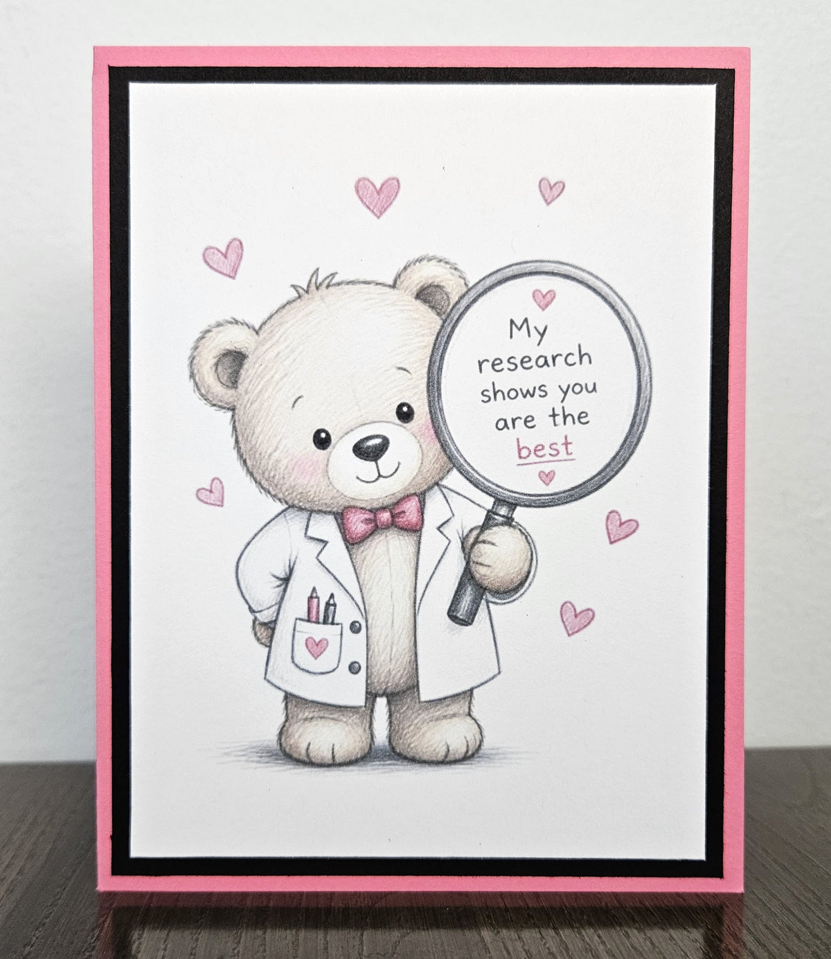 Standing front of a handmade appreciation card with a scientist teddy bear holding a magnifying glass reading My research shows you are the best, framed in pink and black.