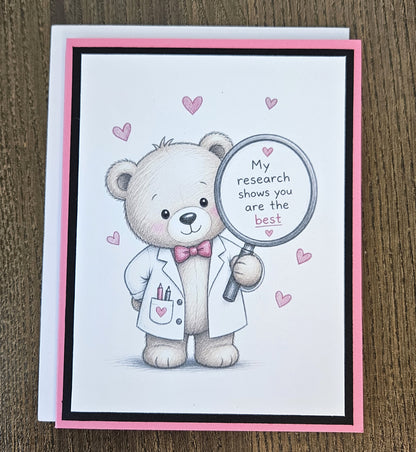 Pink and black scientist teddy bear appreciation card with a white envelope on a wooden surface.