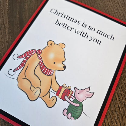 Angled close-up of Pooh in a red striped scarf and Piglet holding a wrapped Christmas present on a black-matted red card.