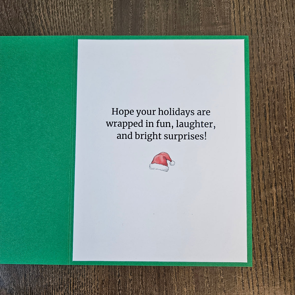 Printed inside of a green Christmas card reading Hope your holidays are wrapped in fun, laughter, and bright surprises, with a small Santa hat below.