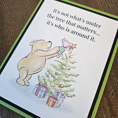 Angled close-up of Pooh and Piglet decorating a Christmas tree beneath the words It's not what's under the tree that matters... it's who is around it.