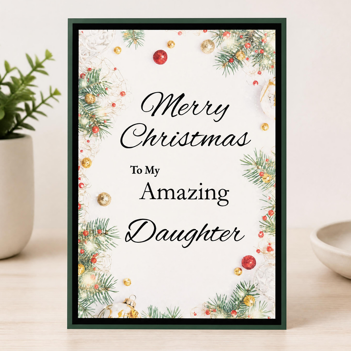 A rectangular Christmas greeting card standing upright on a dark wooden surface against a light gray wall. The card has a dark green border and reads 'Merry Christmas To My Amazing Daughter' in black script and serif text, surrounded by holiday pine branches, red berries, and gold ornaments.