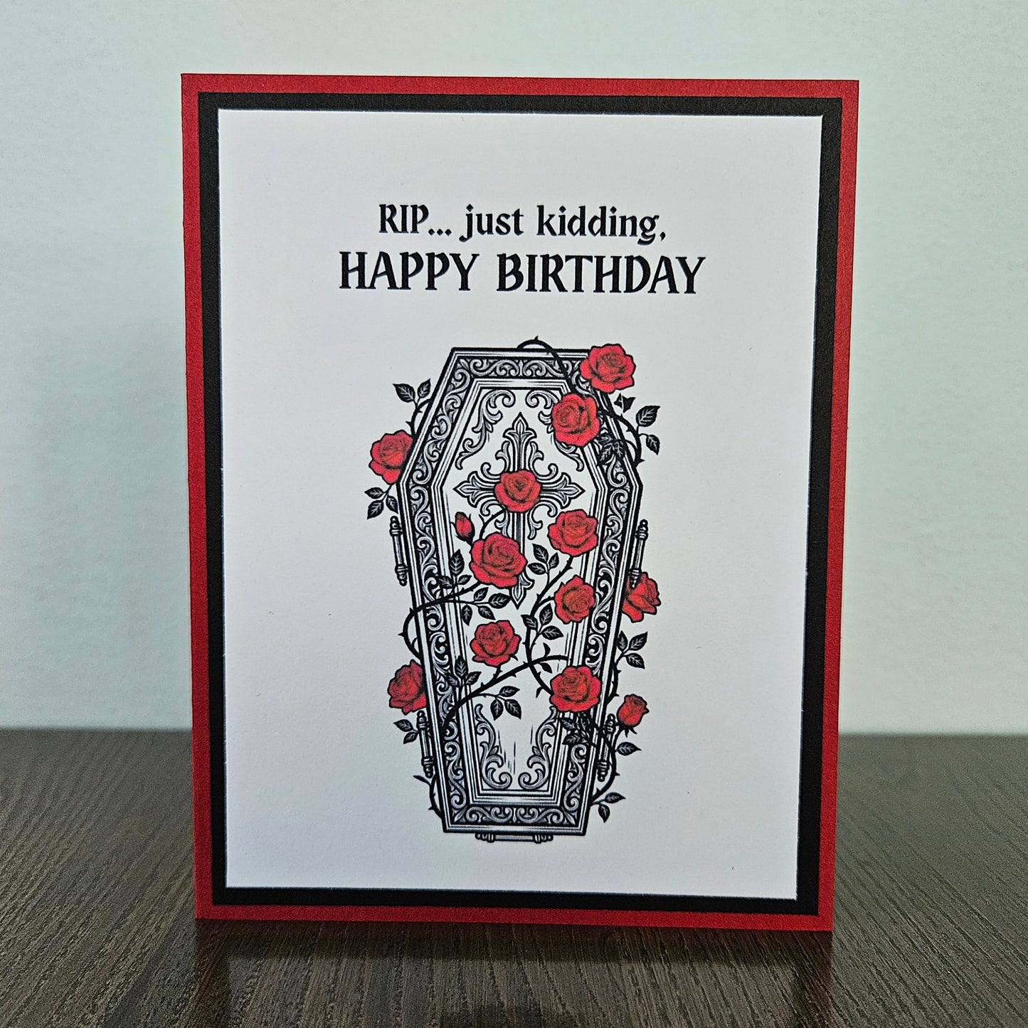 Front of a handmade A2 gothic birthday card featuring a layered design of red, black, and white cardstock. Above an ornate illustration of a coffin covered in red roses and vines is the snarky sentiment: RIP... just kidding, HAPPY BIRTHDAY.