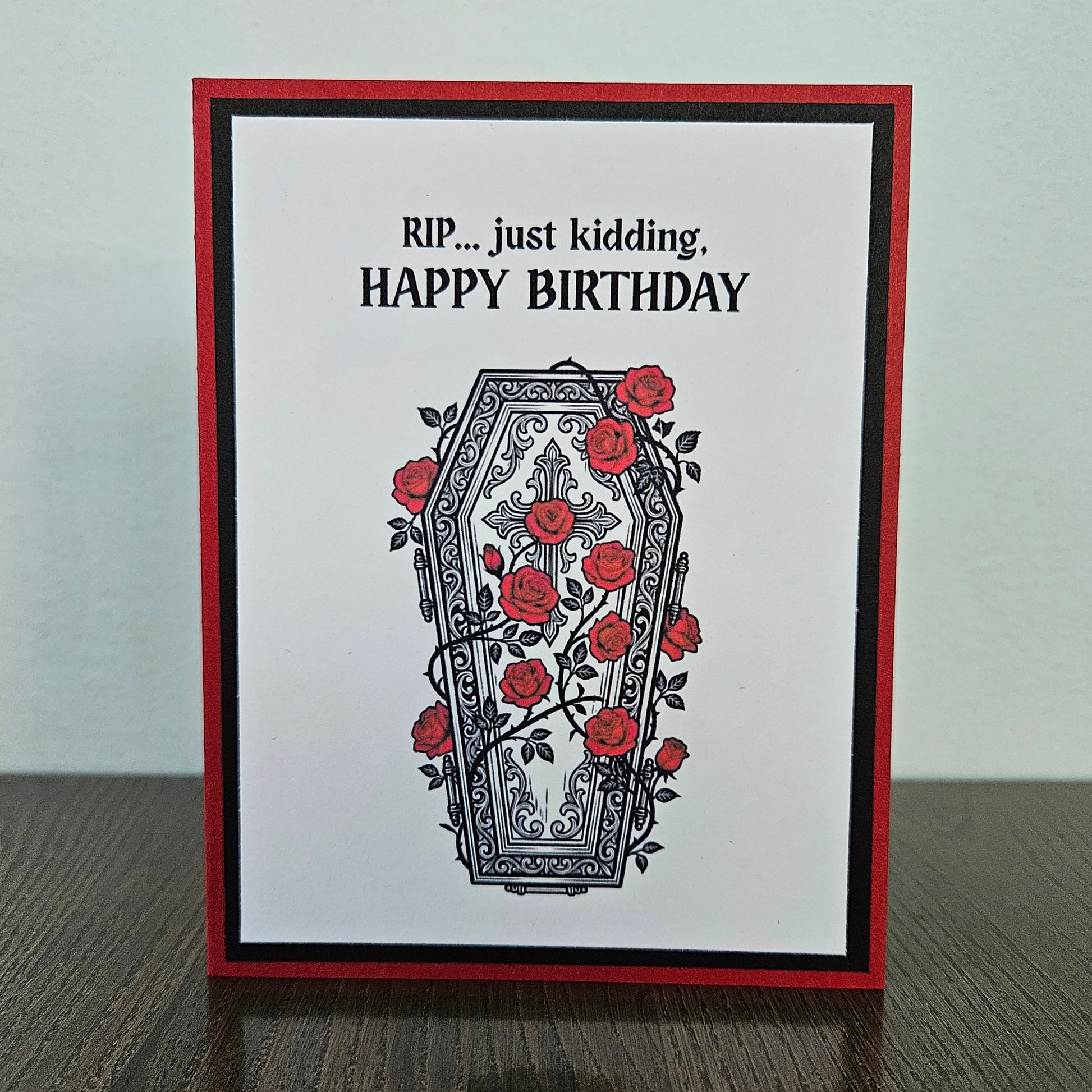 Front of a handmade A2 gothic birthday card featuring a layered design of red, black, and white cardstock. Above an ornate illustration of a coffin covered in red roses and vines is the snarky sentiment: RIP... just kidding, HAPPY BIRTHDAY.