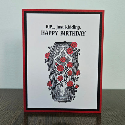Front of a handmade A2 gothic birthday card featuring a layered design of red, black, and white cardstock. Above an ornate illustration of a coffin covered in red roses and vines is the snarky sentiment: RIP... just kidding, HAPPY BIRTHDAY.