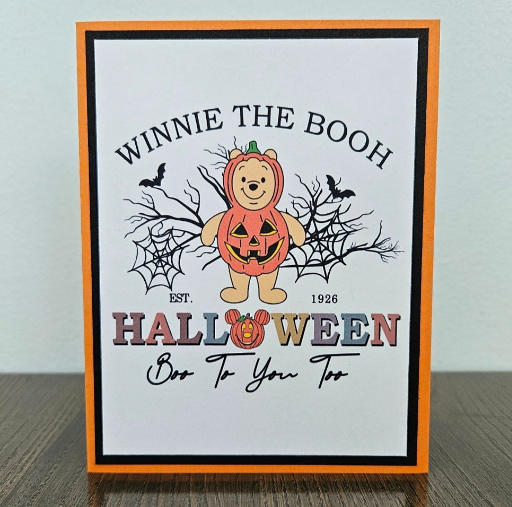 Front of a handmade A2 Halloween card featuring Winnie the Pooh in a pumpkin costume surrounded by spider webs and bats framed in black and orange cardstock with text reading Winnie The Booh Halloween Boo To You Too.