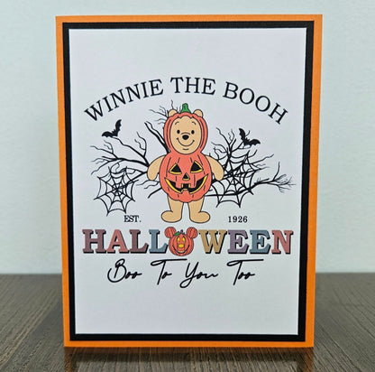 Front of a handmade A2 Halloween card featuring Winnie the Pooh in a pumpkin costume surrounded by spider webs and bats framed in black and orange cardstock with text reading Winnie The Booh Halloween Boo To You Too.