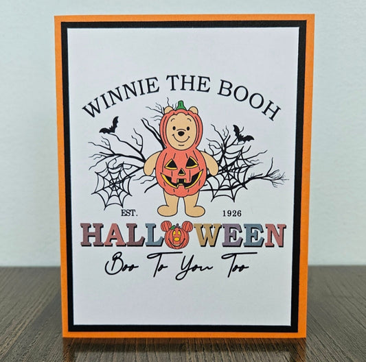 Front of a handmade A2 Halloween card featuring Winnie the Pooh in a pumpkin costume surrounded by spider webs and bats framed in black and orange cardstock with text reading Winnie The Booh Halloween Boo To You Too.
